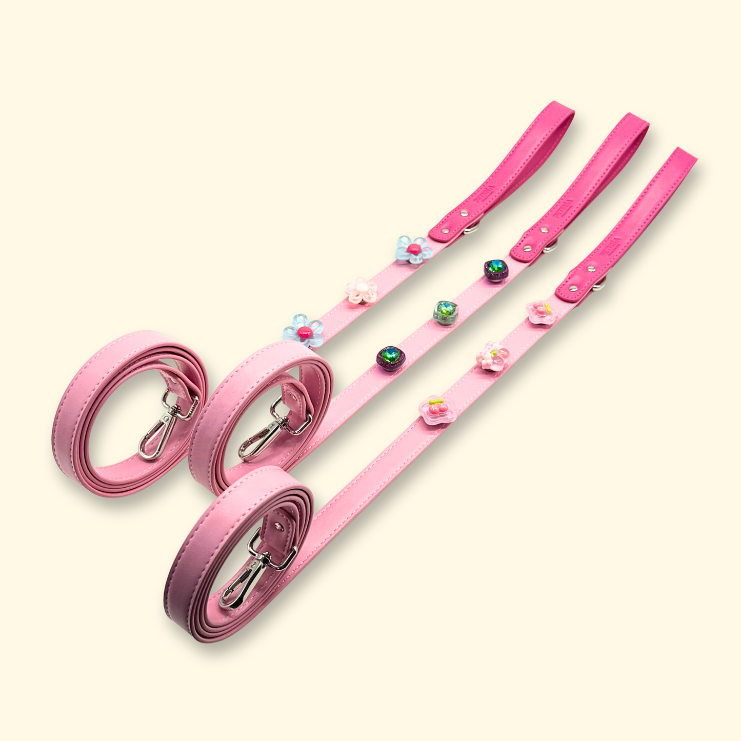 Three blush pink dog leashes laid side by side, each decorated with colorful interchangeable charms including flowers, hearts, and food shapes, on a light beige background.