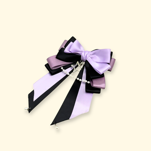 Close-up of Petiria purple and black layered ribbon bow collar charm with pearl chain, showing detailed ribbons and elegant design on beige background
